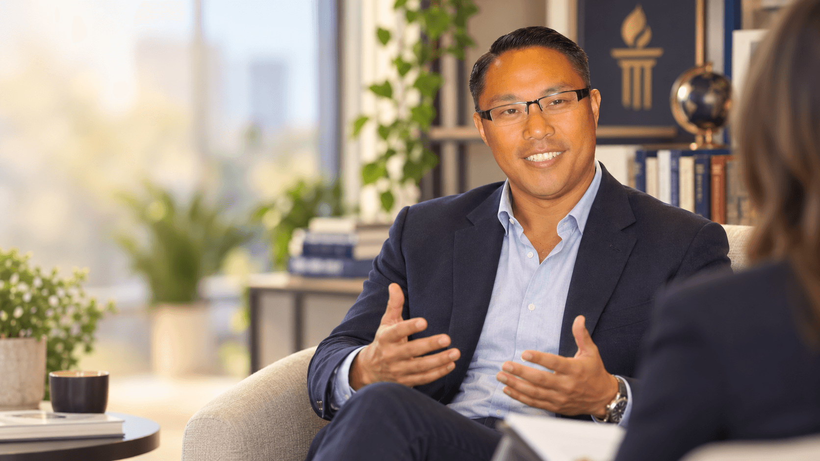 A man in a navy blazer smiling and gesturing while talking with someone in an office, in front of a bookshelf